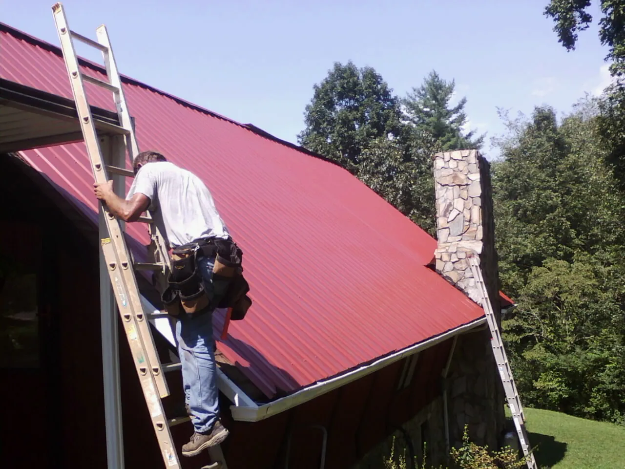 Roofer on a ladder beside a red metal roof and stone chimney — Soffit & Fascia Repair by Roofing Brothers in Osterburg